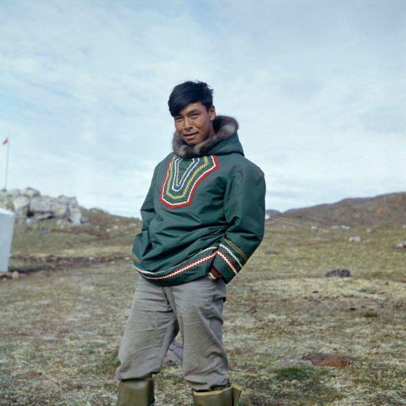 A person stands outdoors in a barren, grassy arctic landscape under a cloudy sky. They are wearing a dark green traditional Inuit parka with fur-lined hood and colorful embroidered patterns in red, yellow, blue, and white around the chest and hem. The person has short black hair, is smiling slightly, and has hands in pockets. They are dressed in light gray pants and green boots. Rocky formations and a flagpole are visible in the background.