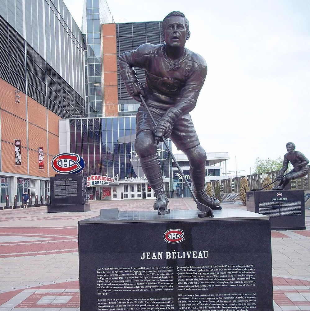 The photo shows a bronze statue of Jean Béliveau in a dynamic skating pose, wearing a Montreal Canadiens jersey, holding a hockey stick, and positioned on a black pedestal with the Canadiens logo and his name 