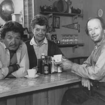 Three people sitting at a counter in a cozy diner, with cups of coffee and a bottle on the table. The background features shelves with various kitchen items and decorations.