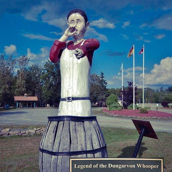 A large wooden barrel with a carved figure of a man wearing a red shirt and white apron, holding a hand to his mouth as if shouting, stands outdoors. The scene features a clear blue sky with scattered clouds, green trees, and three flagpoles with flags, including a Canadian flag. A signpost nearby reads "Legend of the Dungarvon Whooper," detailing a 19th-century tale of a young cook named Ryan who was murdered in a lumber camp near the Dungarvon River, followed by eerie whooping sounds and screams heard for years. The photo includes a link to a website about the legend.