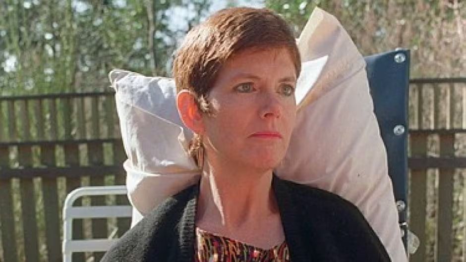 This photo shows an individual seated outdoors. Here's a detailed description:

Subject: The person has short, light brown hair and is wearing a patterned top with a dark cardigan or jacket over it.
Pose: They are sitting in a relaxed manner, with their head resting against a large white pillow that is attached to a dark-colored chair, possibly a lounge or deck chair.
Setting: The background features a wooden fence and greenery, suggesting the photo was taken in a garden or backyard setting. The lighting indicates it might be daytime with natural light illuminating the scene.