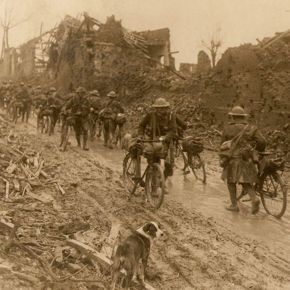 A sepia-toned historical photo of World War I soldiers walking and pushing bicycles along a muddy, debris-strewn path through a destroyed village. Ruined buildings and rubble line both sides. The men wear helmets and greatcoats, some carrying rifles. A dog walks in the foreground.