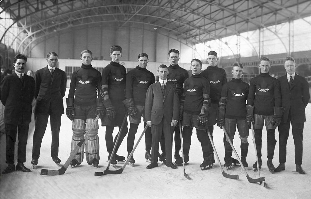 A black-and-white photo of a Canadian hockey team from the early 20th century, standing on an indoor ice rink. Players wear "CANADA" jerseys, hold hockey sticks, and have skates on. Two men in suits stand among them. The rink has an arched ceiling with exposed beams.
