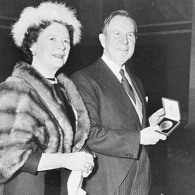 A black-and-white photo of a man and a woman standing together. The woman is wearing a fur stole and a feathered hat, holding a handkerchief. The man is dressed in a suit and tie, holding an open box with a medal inside. There is an additional box on the table in front of them.