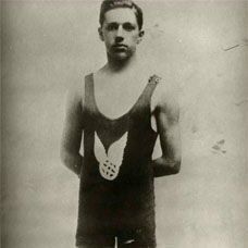### Alt Text for the Photo

A black-and-white vintage portrait photograph of a young man in his late teens or early twenties, standing frontally with hands clasped behind his back in a formal pose. He has a slender, athletic build, short dark hair neatly combed, and a serious, direct gaze at the camera. He wears a dark one-piece tank-style swimsuit with thin shoulder straps, featuring a prominent white emblem on the chest depicting wings flanking a circular medallion or wheel-like design. His legs are bare, and he stands barefoot on a plain studio background. The image has a sepia-toned, early 20th-century aesthetic, with a faint signature or mark in the lower right corner.