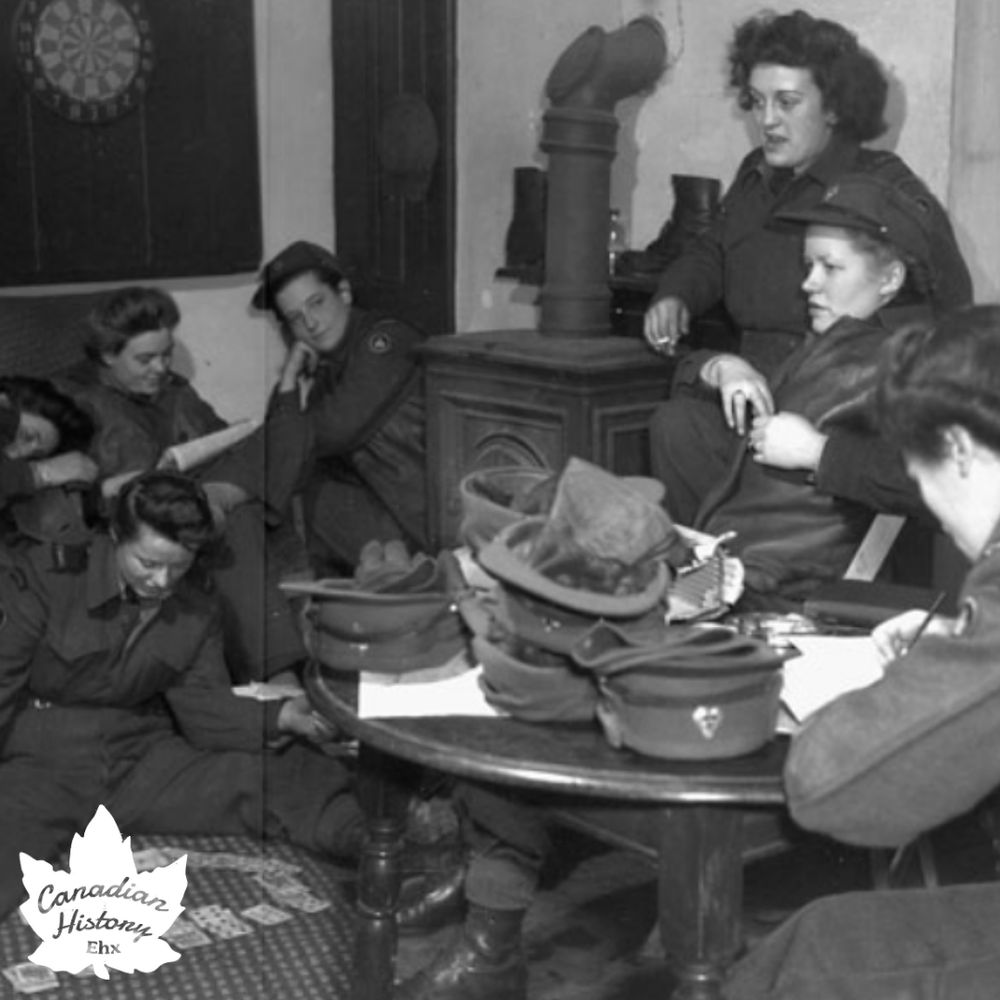 A group of people in military uniforms, likely from the mid-20th century, are gathered in a room. They’re sitting around a table with hats and papers, near a wood stove and a dartboard. Some are writing or reading, while others converse.