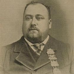 A sepia-toned photograph of a large man with a beard, wearing a formal suit, tie, and a vest. He has several medals pinned to his left side, and he is posed against a plain background.