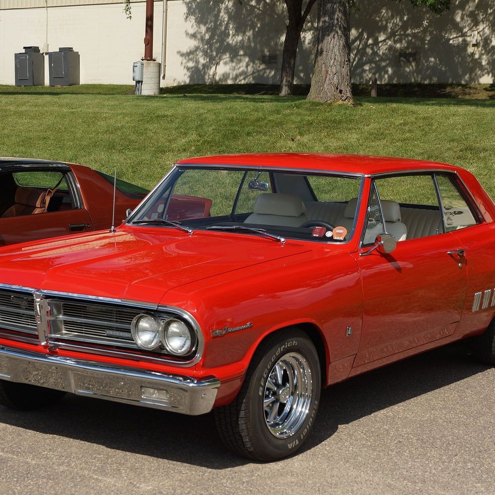 A bright red mid-1960s two-door hardtop coupe parked on pavement at an outdoor car show on a sunny day. The car features a glossy red paint finish, chrome front and rear bumpers, dual round headlights, a horizontal bar grille, chrome Cragar-style wheels with BFGoodrich raised white letter tires, white interior visible through the windows, and "Beaumont" script on the front fender. A portion of another red classic car is visible to the left, with green grass, trees, and a building in the background.