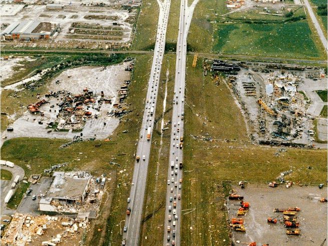 The photo is an aerial view of the Sherwood Park Freeway in Edmonton, Alberta, showing heavy traffic congestion on the multi-lane highway post-disaster. Extensive destruction is visible on both sides, including demolished industrial buildings, scattered debris fields, overturned vehicles and heavy equipment, and ravaged grassy areas adjacent to the road. 