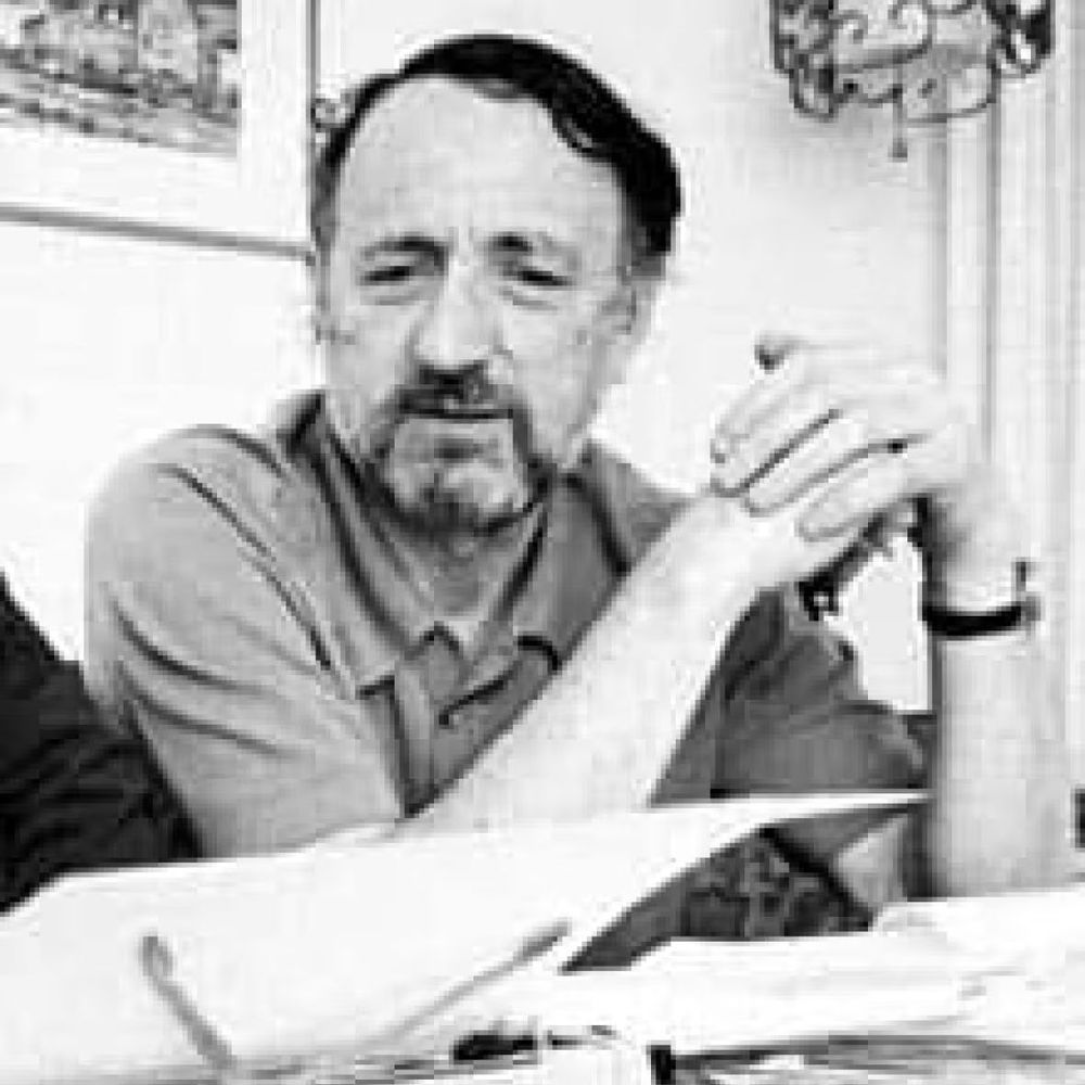 A black-and-white photo of a man with a mustache and short hair, sitting with his arms crossed and resting on a table covered with papers. He is wearing a short-sleeved shirt and a wristwatch. In the background, there is a framed picture on the wall and a decorative wall sconce.