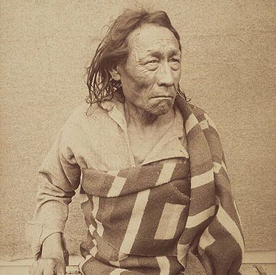 The photograph depicts an elderly individual seated, wrapped in a patterned blanket with a checkered design in shades of brown and white. The person is wearing a loose, long-sleeved shirt and has long, dark hair. Their hands are visible, with one holding a small object and the other resting on a chain that appears to be attached to their wrist, suggesting restraint. The background is plain and neutral, and the image has a sepia tone, indicating it may be historical.
