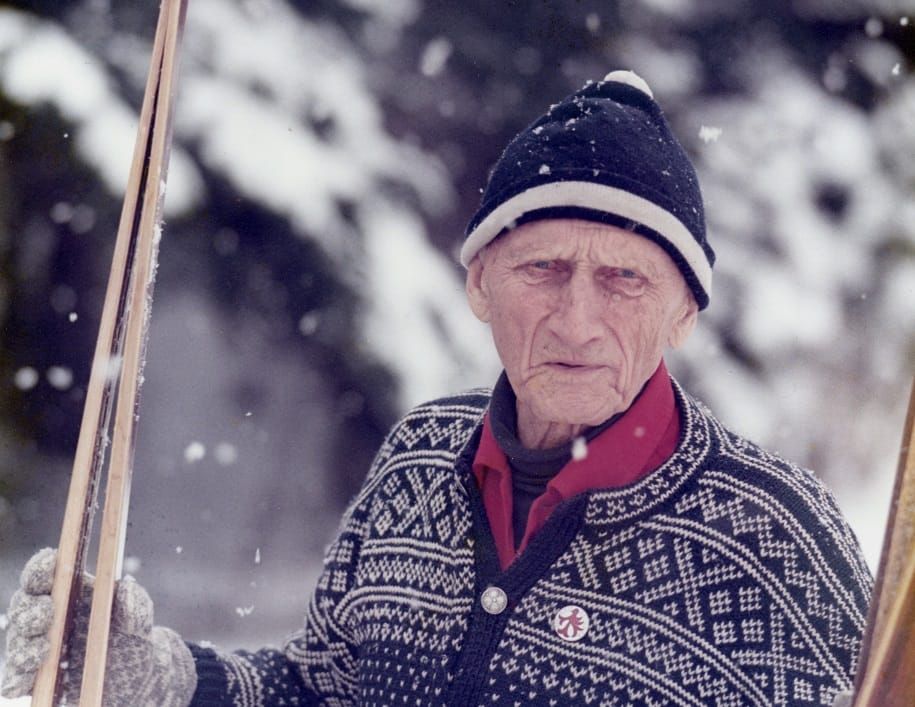 The photo depicts an individual dressed in winter attire, standing outdoors in a snowy environment. They are wearing a patterned sweater with a design typical of traditional Scandinavian or Nordic knitwear, featuring intricate white patterns on a dark background. Underneath the sweater, they have on a red shirt. The person is also wearing a dark knit cap with a white band and a pom-pom on top. They are holding ski poles, suggesting they might be engaged in cross-country skiing. Snow is falling, and the background is filled with snow-covered trees, indicating a cold, wintery setting.