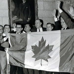 A black-and-white photo of a group of men in suits standing indoors, clapping and raising their arms in celebration while holding a large Canadian flag with a central maple leaf. A framed portrait hangs on the wall behind them.