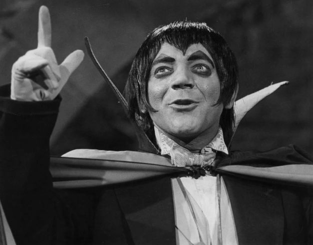 The photo depicts a person dressed as a vampire, complete with exaggerated makeup and costume. The individual has pale skin, dark arched eyebrows, and is wearing a black cape with a high collar, typical of classic vampire attire. They are also making a hand gesture with two fingers extended, which could be interpreted as a playful or dramatic gesture. 