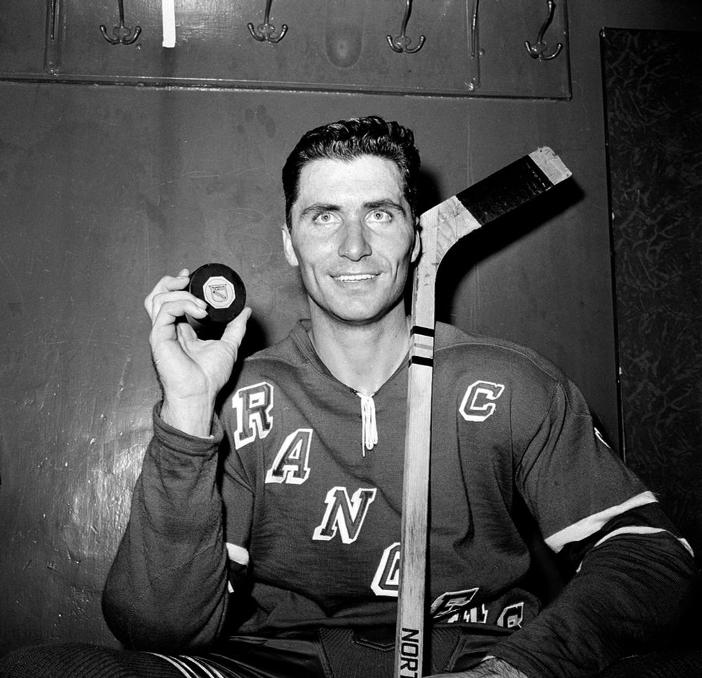 The black-and-white photo shows a hockey player wearing a jersey with "RANGERS" written across the chest and a captain's "C" on the sleeve. He is holding a hockey puck in one hand and has a hockey stick resting on his shoulder. The setting appears to be a locker room, with hooks visible on the wall behind him.
