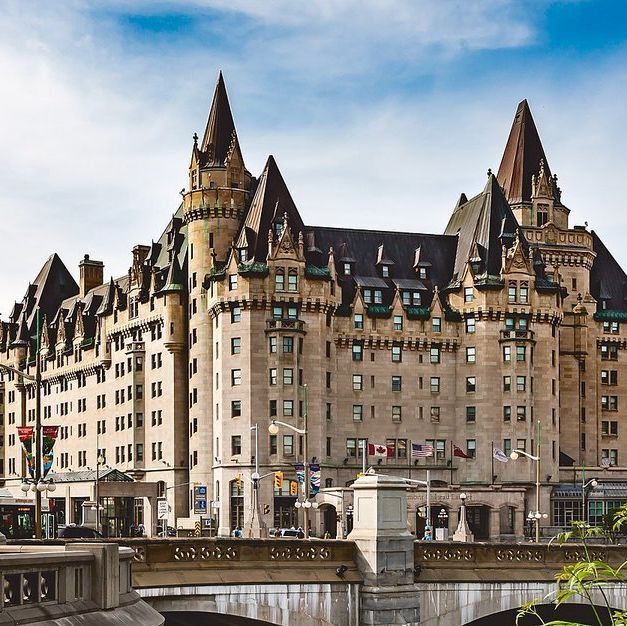 A grand, historic hotel with a Châteauesque architectural style, featuring steep, pointed roofs, dormer windows, and ornate detailing. The building is multi-storied with a light beige facade and dark green roofs, situated alongside a stone bridge with decorative railings. The scene is set under a partly cloudy sky, with some greenery and urban elements visible in the foreground.