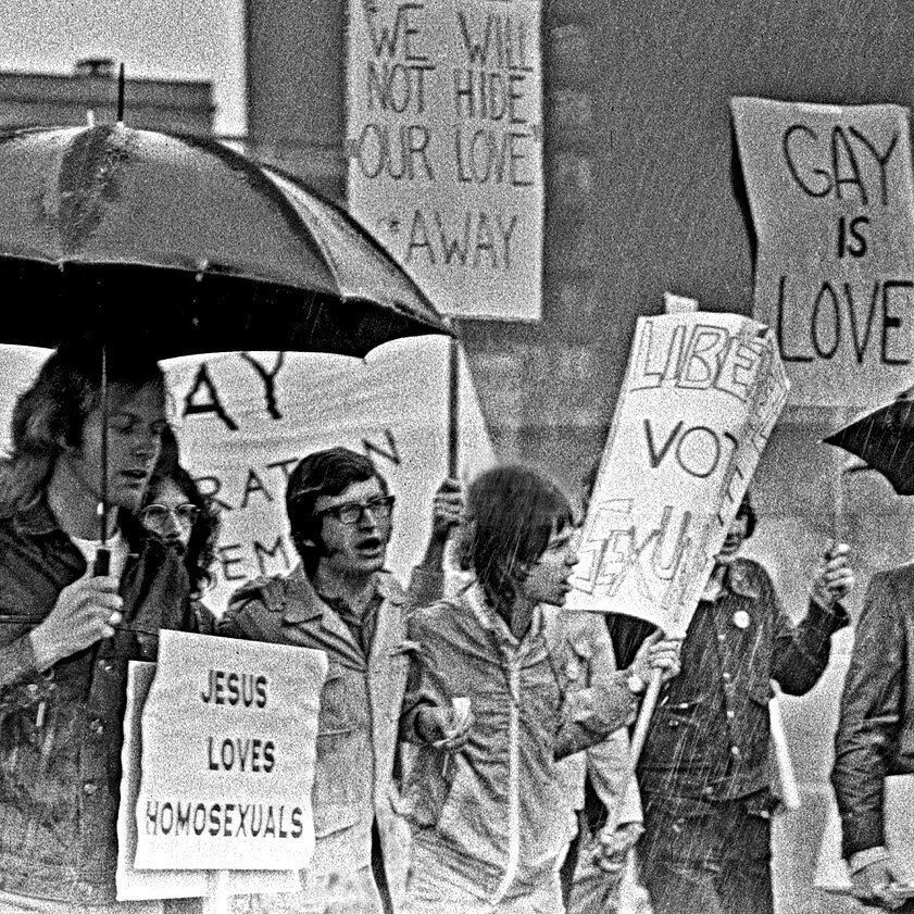 A black-and-white photo of a group of people marching in the rain under umbrellas, holding signs with messages such as "JESUS LOVES HOMOSEXUALS," "WE WILL NOT HIDE OUR LOVE AWAY," "GAY IS LOVE," and "LIBERATE YOU." The scene appears to be a protest or rally, with participants dressed in 1970s-style clothing.