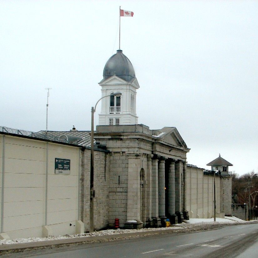 A photo of a large, historic stone building with a domed tower and columns, topped with a Canadian flag. The structure is surrounded by high walls and a watchtower, with a sign on the wall reading "Guides." The scene is set on a cold day with snow on the ground and a gray sky, and a road runs in front of the building.