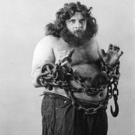 A shirtless person with a long beard and curly hair stands with their hands raised, wrapped in heavy chains with hooks around their torso and arms, wearing dark pants.