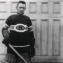 A black-and-white photograph of a hockey goalie standing in front of a paneled wooden door. The goalie is wearing a dark sweater with a prominent "CH" logo on the chest, large padded leg guards, gloves, and holding a hockey stick vertically. The image has a vintage style, likely from the early to mid-20th century.