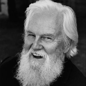 A black-and-white portrait of an elderly man with long white hair and a full white beard, smiling warmly. He wears a dark suit jacket, white shirt, striped tie, and a small flower pin on his lapel. The background is softly blurred outdoor greenery.