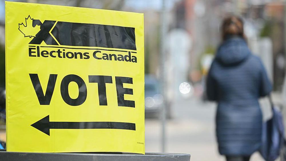 A yellow Elections Canada sign with a red maple leaf and black "X" logo reads "VOTE" with a left arrow. In the blurred background, a person in a blue coat walks on a street.