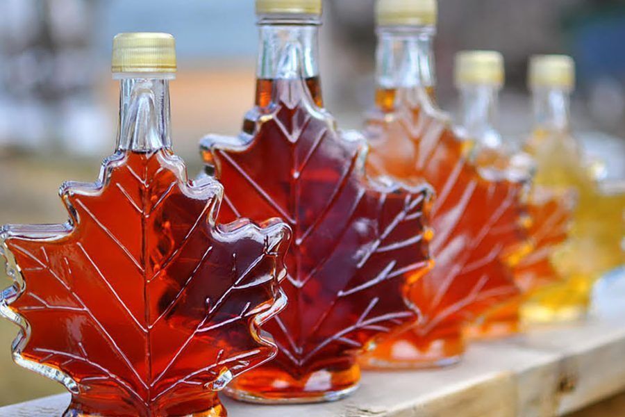 A row of small glass bottles shaped like maple leaves, filled with maple syrup in varying shades from deep amber to light golden. The bottles have gold-colored caps and are lined up on a wooden surface outdoors, with a blurred natural background. The syrup inside glows warmly in the light, highlighting the detailed leaf veins embossed on the glass.