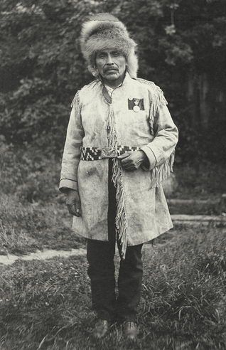 The black-and-white photograph depicts an individual standing outdoors, dressed in traditional attire. The person is wearing a fringed coat with a patterned belt and a fur hat. The background features a natural setting with grass and trees, suggesting a rural or wilderness environment.

