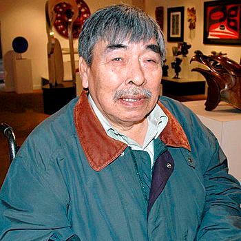 An older Inuit man with gray hair and a mustache sits indoors in an art gallery, smiling gently at the camera. He wears a teal jacket with a brown corduroy collar over a white shirt. Behind him are various sculptures and framed artworks on display, including a large abstract red piece and bronze animal sculptures on white pedestals. The setting is warmly lit.