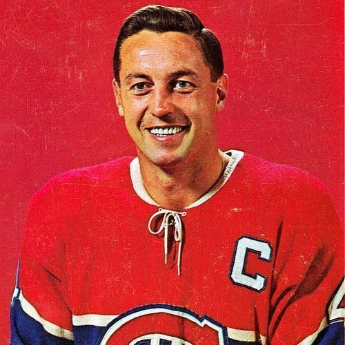 A vintage color photograph of Jean Béliveau, the legendary Montreal Canadiens hockey captain, smiling confidently while seated against a solid red background. He has neatly combed dark hair, sharp features, and wears a classic red Canadiens jersey with blue and white stripes, the team's "CHC" logo on the chest, and a white captain's "C" patch on the left shoulder. The image has a slightly faded, retro filter typical of 1960s hockey trading cards.