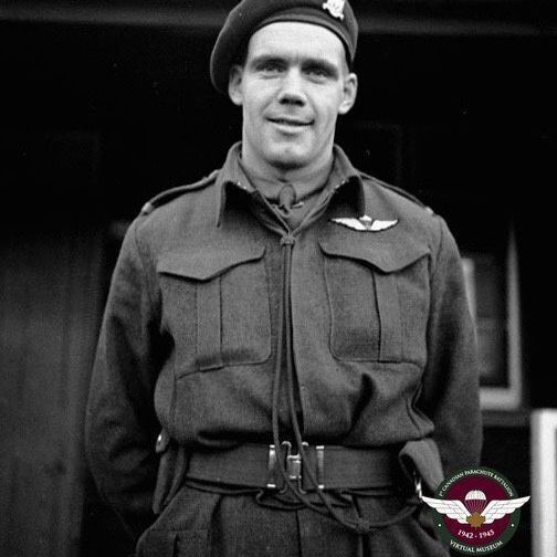Black and white vintage photograph of a young man in British Army paratrooper uniform, standing with hands behind his back and smiling slightly at the camera. He wears a dark beret with a cap badge tilted to the left, a battledress jacket with parachute wings on the left chest, shoulder epaulettes, a belt with buckle, and a lanyard. The background shows a dark wooden building with a doorway. A small circular logo in the bottom right reads "Canadian Parachute Battalion 1942–1945 Virtual Museum" with a winged parachute emblem.