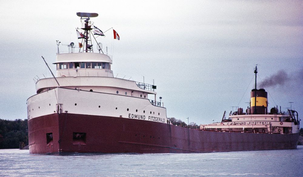 The image shows a large lake freighter ship named "Edmund Fitzgerald" sailing on a body of water. The ship has a white superstructure with a red hull and is flying a flag from its mast. The sky is overcast, and there is a plume of smoke coming from the ship's smokestack, indicating it is in operation. The ship is well-known due to its tragic sinking in a storm on Lake Superior in 1975, which inspired the song "The Wreck of the Edmund Fitzgerald" by Gordon Lightfoot.