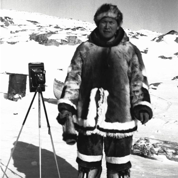 A person wearing a thick fur coat and boots standing in a snowy landscape with mountains in the background, next to a tripod-mounted camera, with a small boat and other equipment visible nearby, in a black-and-white image.