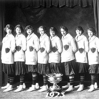 A black-and-white photograph of a women's basketball team from 1923, standing in a line. They are wearing matching uniforms with dark skirts, light-colored tops with dark lettering, and knee-high stockings with stripes. One player on the left is holding a basketball, and a trophy is placed on the floor in front of the team. The background features a draped curtain. The year "1923" is visible at the bottom of the image.