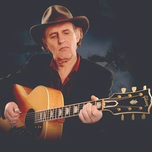 A person wearing a brown hat and a dark suit with a red shirt, playing an acoustic guitar with a visible Gibson logo, set against a dark background with a subtle outdoor scene.