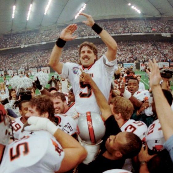 A football player wearing a white jersey with the number 5 is hoisted triumphantly on his teammates' shoulders on the field of a packed indoor stadium. He has a mustache, raises both arms high in celebration, and smiles widely as the crowd cheers under bright stadium lights. Teammates in white jerseys with orange and black accents look up at him joyfully while photographers capture the moment.