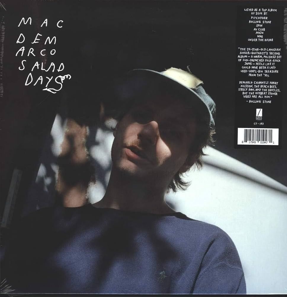 The image shows the back cover of Mac DeMarco's "Salad Days" vinyl album. Handwritten white text in the top-left reads "M A C D E M A R C O S A L A D D A Y S" in a scribbled style. Below is a close-up of a young man (likely DeMarco) looking down thoughtfully, face shadowed by sunlight through leaves. He wears a backward blue cap, dark blue sweater, and has medium brown hair. The background is a plain white surface, giving a chill vibe.

