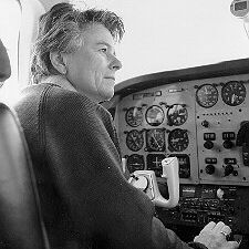Black-and-white photograph of an older person with short, wavy hair, wearing gloves, seated in the left pilot seat of a small airplane cockpit. They are looking out the side window, hands on the control yoke. The instrument panel is filled with analog gauges and dials typical of a light aircraft from the mid-20th century. Bright daylight streams in through the windshield and side window.