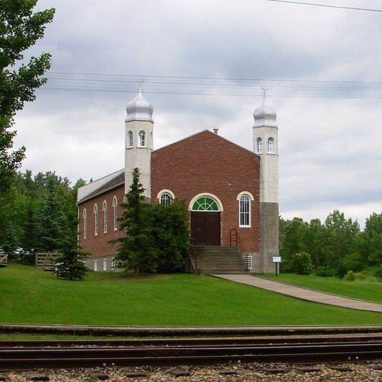 A brick mosque with two white towers, featuring arched windows and a central green arched window above the entrance. The mosque is surrounded by green grass, trees, and a cloudy sky, with railroad tracks in the foreground.