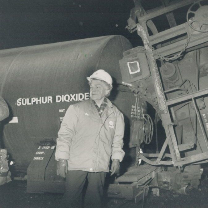 A black-and-white nighttime photo shows a worker in a white hard hat and light jacket standing in front of a derailed, tilted tank car labeled "SULPHUR DIOXIDE" on the left and a damaged structural frame with chains and text on the right. Debris litters the ground in the dark background.