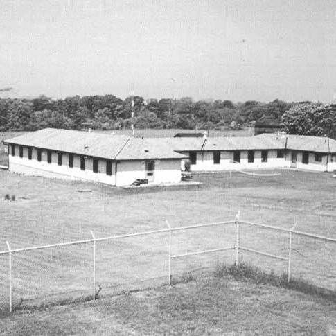 Black and white photograph showing a fenced-in open yard with patchy grass and dirt. In the foreground is a chain-link fence topped with barbed wire. Behind the fence are several single-story rectangular buildings with flat roofs and multiple windows along the sides, arranged in a U-shape around the yard. The buildings appear to be simple, utilitarian structures with light-colored walls and darker roofs. In the background are trees and distant structures under a clear sky.