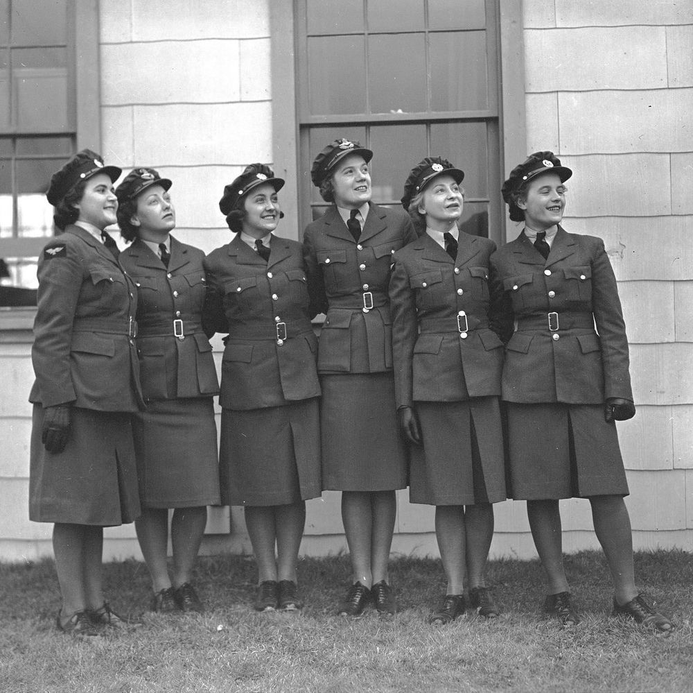 Six women in military uniforms stand in a row in front of a building with large windows, wearing caps, jackets with buttons, skirts, and gloves, with grass at their feet.