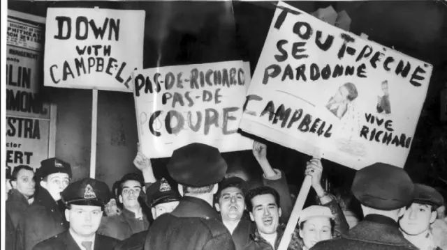 The photo is a black-and-white image capturing a protest scene, likely from the mid-20th century, featuring a group of people holding signs and chanting. The crowd includes several individuals, some wearing hats typical of the era, and a few police officers in uniforms with peaked caps. Prominent signs include one in English reading "DOWN WITH CAMPBELL PAS-DE COUPE" and another in French stating "TOUT SE TUE PRÊCHE PARDONNÉE CAMPBELL VIVE RICHARD," with a drawing of a person on a cross. The background shows additional posters on a wall, suggesting an urban setting, possibly outside a public building or arena. The mood appears tense, with the protesters expressing strong opposition, likely related to a political or social issue involving someone named Campbell, possibly tied to a hockey-related controversy given the mention of "Richard," which could refer to Maurice "Rocket" Richard, a famous Montreal Canadiens player.
