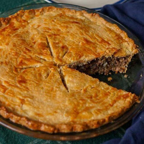A golden-brown baked pie in a clear glass pie dish, with a flaky crust featuring decorative slits on top forming a star-like pattern. One slice has been removed, revealing a dark meat filling inside. The pie sits on a green plaid tablecloth with a dark blue napkin partially visible beside it. The crust appears crisp and glossy, likely brushed with egg wash.