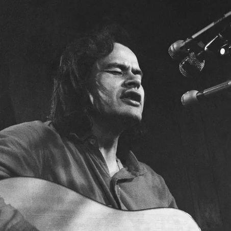 Black and white photograph of Willie Dunn, a man with long dark hair and a beard, eyes closed and mouth open wide as if singing passionately, while playing an acoustic guitar. He is wearing a long-sleeved shirt, positioned in front of multiple microphones suspended nearby, against a dark background.