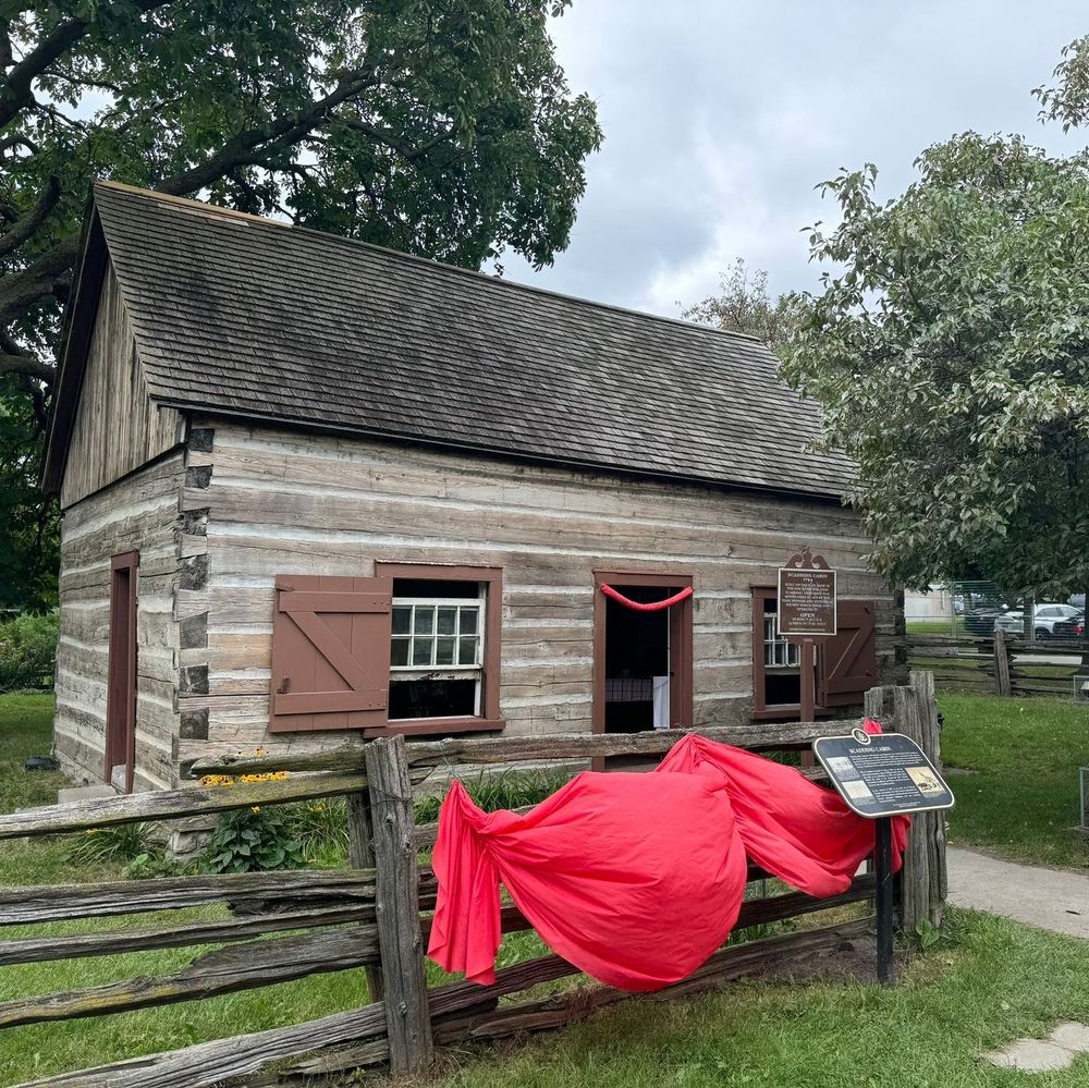 A color photograph of a small historic log cabin with weathered wooden walls and a shingled gable roof, featuring a front door flanked by two small multi-pane windows with brown shutters and one open window. Red fabric drapes are tied to the wooden split-rail fence in the foreground, along with informational plaques on the fence and cabin wall reading "Scadding Cabin" and details about its history as Toronto's oldest house, built in 1794, open to the public. Large green trees frame the scene under a cloudy sky, with a grassy area, flowers, sidewalk, and distant parked cars visible.