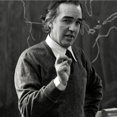 The black-and-white photo shows filmmaker Colin Low standing in front of a chalkboard with a map outline. He is wearing a sweater over a collared shirt and tie, gesturing with his right hand while his left hand rests on a table.