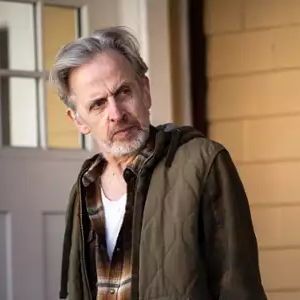 A middle-aged man with gray hair and a short beard, wearing a green quilted jacket over a plaid shirt and white undershirt, stands in front of a beige wall and white door, gazing intently to the side with a serious expression.