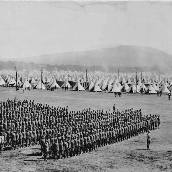 Black-and-white photograph showing a large formation of soldiers standing in aligned rows on a flat open field, with several individuals positioned in front of the group. In the background, numerous conical tents are arranged in rows extending into the distance, accompanied by scattered small groups of people, a few vehicles including a car and horse-drawn carts, utility poles, and hazy hills or mountains on the horizon.