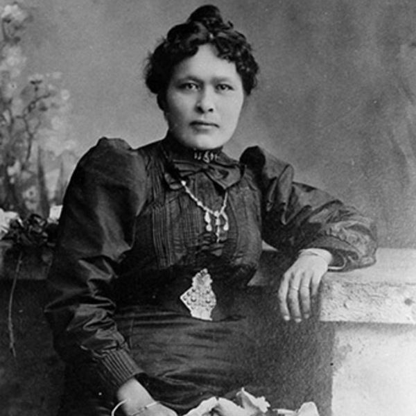 A black-and-white photograph of a woman with her hair styled in an updo, wearing a dark, long-sleeved dress with puffed sleeves, a high collar, and a bow at the neck. She has a necklace and a decorative brooch, and is holding a bouquet of flowers while resting her arm on a surface.